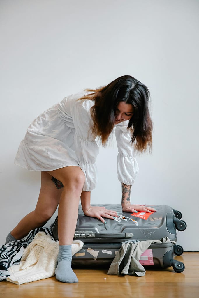 A woman struggles to close an overpacked suitcase, with belongings scattered around, showing how not to pack for surgery abroad.